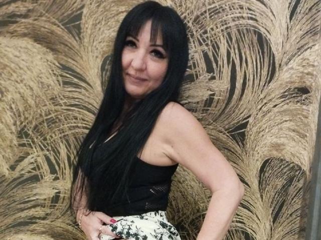 SusanLaNicex with long black hair, smiling, wearing a black top and white floral skirt, standing in front of large, beige, feathery wall, showing her ass with a hand on it.