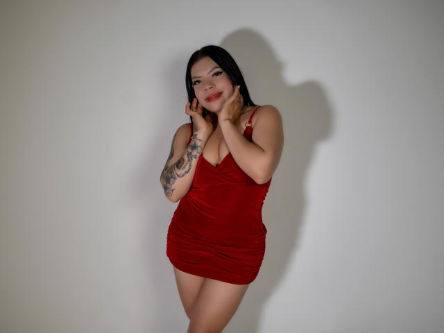 AdrianaPaul in a red velvet dress, hands on her face, showing off her tattoos, and her medium-sized tits, with a sexy smile, standing against a plain white wall.
