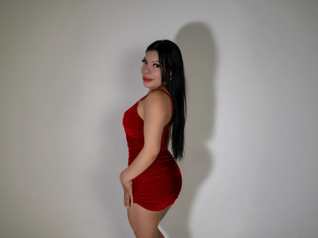 AdrianaPaul, con i suoi lunghi capelli neri, l'abito di velluto rosso e il rossetto rosso, che mette in mostra i suoi fianchi prosperosi e i piccoli seni, è in piedi di lato con un sorriso sicuro..