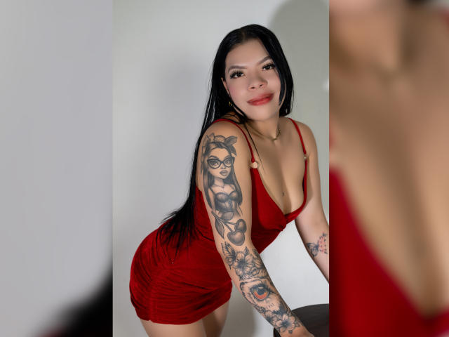 AdrianaPaul in a red dress, leaning forward, showing off tattooed arm with glasses girl and other tattoos, long black hair, smiling, red lipstick, small gold necklace, cute.