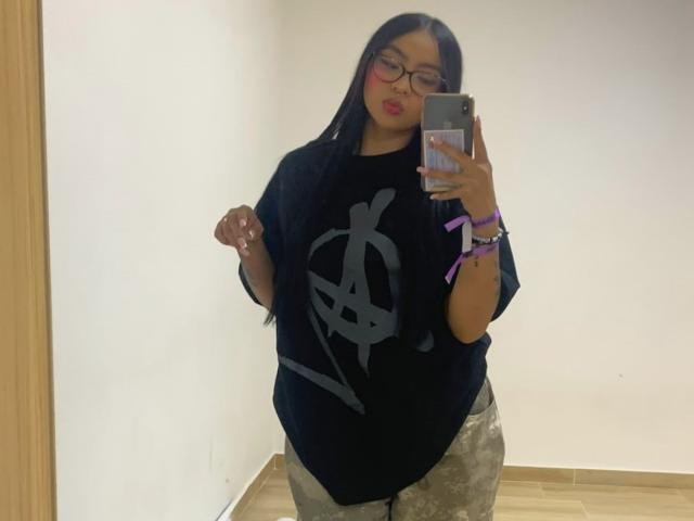 ChloeMoretti with long black hair, glasses, and pink lipstick, wearing a black shirt with a gray graphic, camouflage pants, and purple bracelets, taking a mirror selfie.