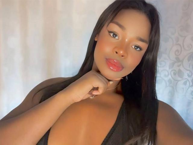 VenusWilly with glossy pink lips, dark straight hair, and brown skin, touching her chin with a hand, wearing a black top, showcasing her large, shiny, bare tits.