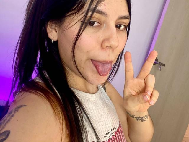 SophiaMarx with black hair in a ponytail, sticking out her tongue, making a peace sign, wearing a white sleeveless top, showing a tattoo on her left arm.