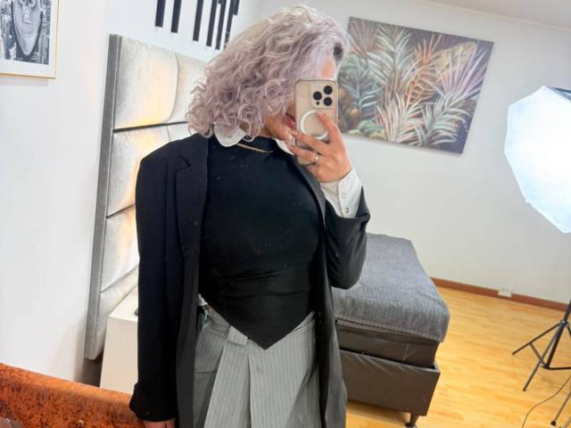 Elena Saenz, con cabello gris rizado, chaqueta negra, camisa blanca, top negro, falda gris a rayas, sosteniendo un teléfono, reflejada en un espejo, suelo de madera, pared blanca, iluminación de estudio..