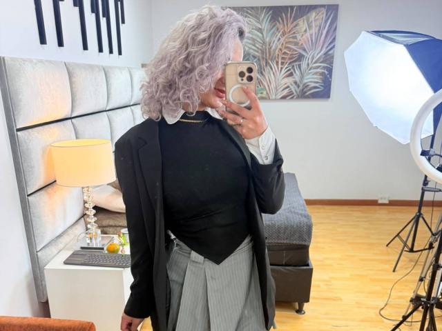 Elena Saenz, con cabello gris rizado, blusa negra, falda gris a rayas finas y chaqueta negra, sosteniendo un teléfono, selfie con espejo, luz de aro, lámpara suave, cama con cabecera gris en el fondo..