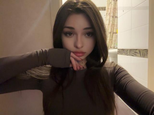 RayleneMazzeo with long brown hair, wearing a gray long-sleeve top, resting her chin on her hand, looking at the camera with a slight pout, in a bathroom.