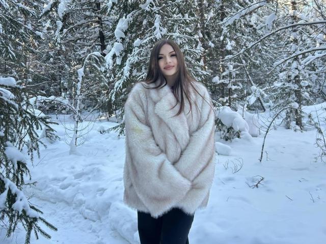 RayleneMazzeo stands in a snowy forest, wearing a fluffy white fur coat, black pants, and a slight smile. She has long brown hair, and her tits are not visible.