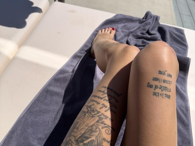 SensualMila's tattooed legs, with her foot on a gray towel, on a white sunbed. She's got ink on her thigh and calf, with red-painted toenails.