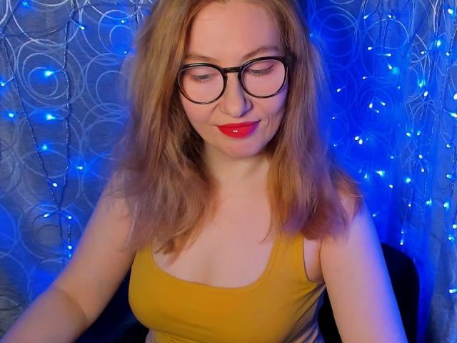 PaulinaSunny with red lipstick, glasses, and yellow tank top, showing small tits, smiling, sitting, and blonde hair, blue light in background.