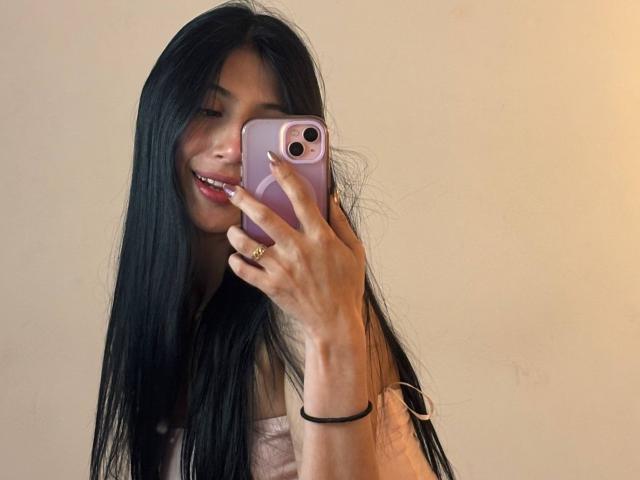 Sophie Beaumont, avec de longs cheveux noirs, souriante, tenant un téléphone rose, montrant son visage et sa main avec une bague, portant un haut rose, sur un fond uni..