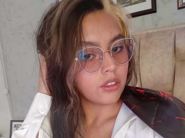 RubyLane has dark and blonde hair, wearing clear glasses, white shirt, and black jacket with red pattern, looking slightly annoyed, hand in hair.