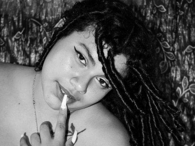 Fotografia in bianco e nero di Victoriaa Jackson con trecce ricci, che si morde l'unghia dipinta di bianco, con lo sguardo rivolto verso l'alto e con l'eyeliner ad ali di farfalla, che indossa una collana, su uno sfondo a motivi..