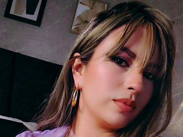 AmiiRosse has light brown hair with bangs, wearing purple and gold earrings, looking at the camera with a slight smile, soft pink lipstick, and subtle makeup.