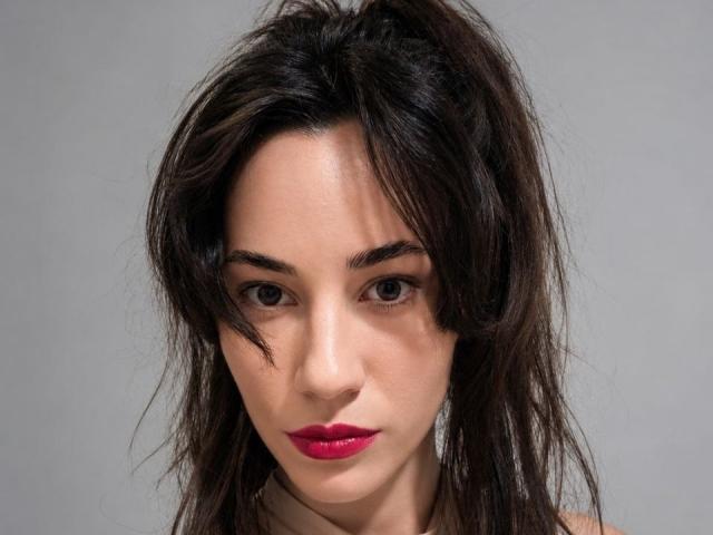 ArianeEsther has messy dark brown hair, intense brown eyes, and bright red lipstick. Her face is centered, showing a serious expression with slightly parted lips.