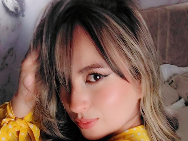 AmiiRosse ha i capelli biondi e neri, indossa una camicia gialla a pois, ha un sorriso carino e un trucco per gli occhi, e si tocca i capelli. La sua pelle è chiara e ha le labbra rosa..
