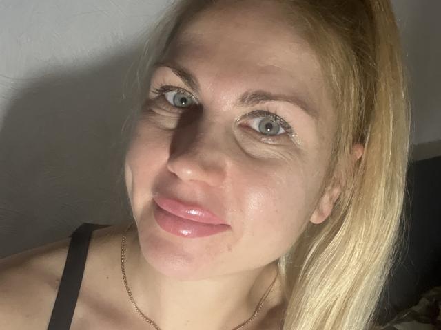 LikaBlossy heeft glanzende, roze lippen, blauwe ogen en blond haar, draagt een zwarte bandeau met een subtiele gouden ketting, en heeft een lichte glimlach op een goed verlichte foto..
