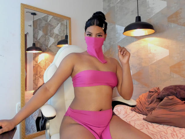 LalilaBrunette ist eine kurvige Frau mit mittelbrauner Haut, die einen pinken Bandeau-Top und passende Unterwäsche trägt, ihr schwarzes Haar ist zu einem Dutt gebunden, sie trägt eine pinke Maske und sitzt auf einem weißen Stuhl..