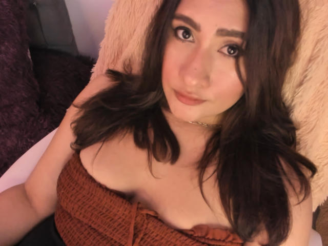 ElyRoss with brown wavy hair, wearing a strapless burnt-orange top, revealing her boobs. She's looking at the camera with a slight smile, lying on a fluffy pillow.