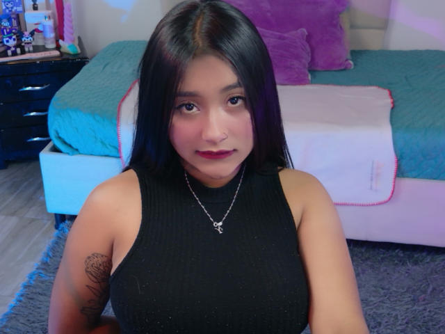 AmmyLewis with long black hair, wearing a black sleeveless top, showing her large tits, tattoo on her right arm, and a silver necklace, smiling in a bedroom.