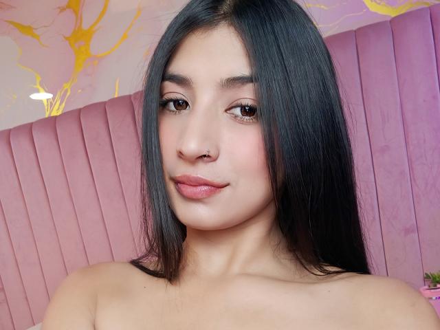 Naia Sanders tiene el cabello negro y lacio, ojos marrones y labios rosados, además de un piercing en la nariz. Está sin ropa, mostrando sus hombros lisos y un ligero bronceado. Su expresión es relajada y linda..