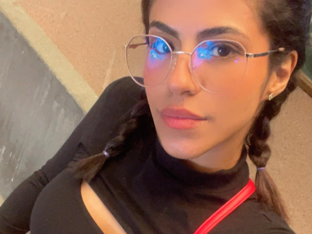 EvaGate with medium brown skin, braided hair, wearing clear glasses, black turtleneck, red strap, and pink lips, showing her tits, looking at camera with a slight smile.