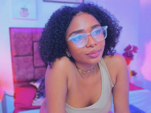 MeganWlliams with curly black hair, clear glasses, tan tank top, gold chain, and nose ring, showing small tits, sitting in a colorful room with pink and blue lighting.