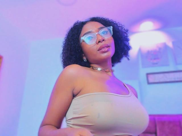 MeganWlliams with curly black hair, wearing glasses, beige tank top, and necklace, showing large, rounded, bare boobs, standing in purple and blue lit room.