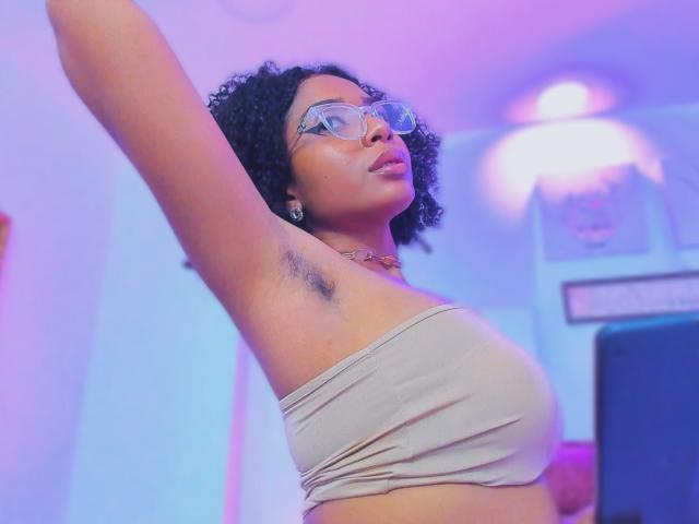 MeganWilliams with curly black hair, wearing clear glasses, beige tube top, exposing armpit, arms raised, looking up, pink and purple lighting, medium brown skin, small gold necklace.