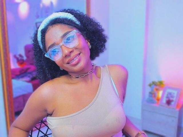 MeganWlliams with curly black hair, wearing clear glasses, white headband, chain necklace, and one-shoulder beige top, smiling at the camera, showing her small tits.