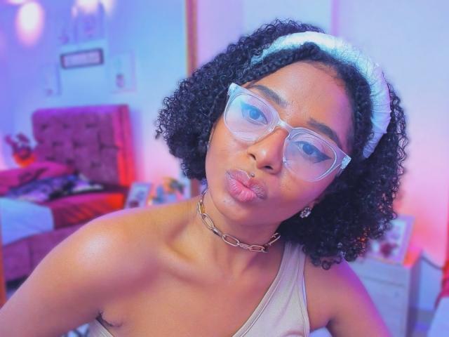 MeganWilliams with curly black hair, clear glasses, white headband, chain necklace, and pouty lips, wearing a gray tank top, showing shiny skin and small brown nipples.