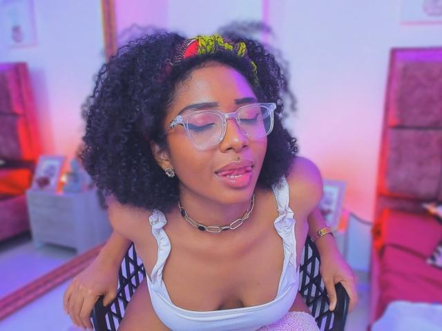 Megan Williams, com cabelo preto encaracolado, óculos de grau, faixa amarela na cabeça, top branco que mostra seus seios grandes, colar de corrente, sentada em uma cadeira preta, língua para fora, iluminação roxa e rosa..