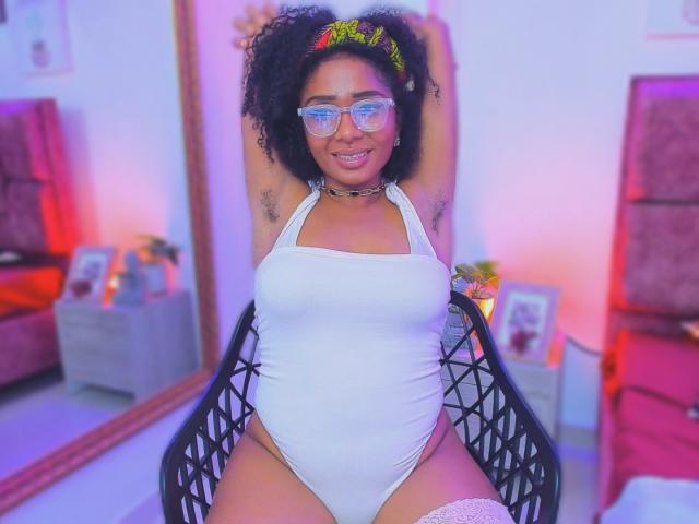 MeganWlliams, dark-skinned with curly hair, wearing glasses, white halter one-piece, arms raised, showing armpit hair, choker, and white thigh-highs, smiling.