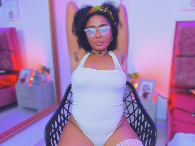 MeganWilliams with curly black hair, wearing glasses, white halter one-piece, and a yellow headband, arms raised, showing armpit hair, sitting in a black chair, legs apart.