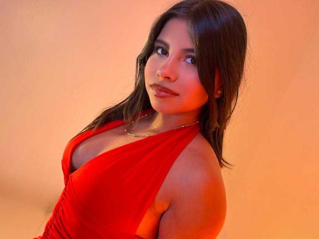 Celeste Morel, con cabello oscuro, vestida con un vestido rojo con un escote pronunciado, mirando a la cámara con una sonrisa sutil, con sus senos ligeramente visibles, sobre un fondo naranja cálido..