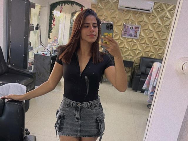 EvaGate with medium tits, black v-neck shirt, and denim skirt, holding phone, standing in room, mirror selfie, brown hair, slight smile, air conditioning above.