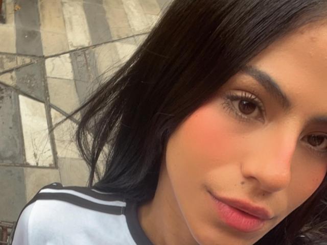 EvaGate has dark brown eyes, a small nose piercing, and a subtle smile. She's wearing a white shirt with black stripes, and her black hair is down.