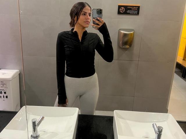 EvaGate with dark brown hair, black long-sleeve shirt, and white pants, taking a mirror selfie in a bathroom, showing her butt in tight pants.