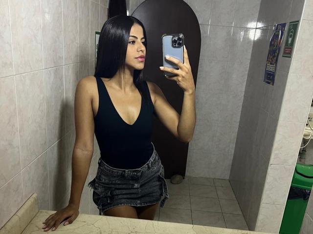 EvaGate with long black hair, black tank top showing her tits, and a denim skirt, taking a mirror selfie with her right hand, in a tiled bathroom.
