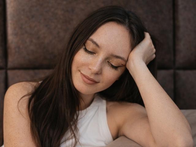 WendyDemmy with long brown hair, freckled face, closed eyes, subtle smile, white sleeveless top, hand in hair, glittery gold eyeliner, brown patterned headboard, relaxed, cute.