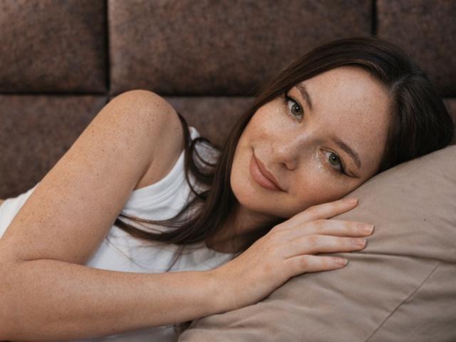 WendyDemmy with freckles, lying on a pillow, wearing a white tank top, smiling softly, brown hair, green eyes, and resting her head on her hand, showing a cute face.