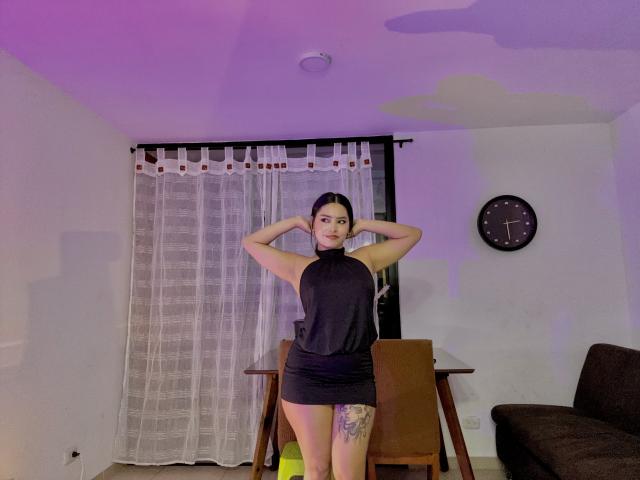 PamelaArabel está em pé, com as mãos atrás da cabeça, vestindo uma blusa preta sem mangas e uma saia curta, mostrando uma tatuagem na parte interna da coxa direita, em um quarto com iluminação roxa..