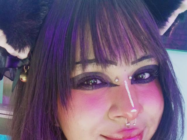 El rostro de Kuroomi con maquillaje morado y rosa, cabello morado oscuro, orejas de gato negras, un aro en la nariz y un piercing en el tabique, y ojos verdes. Está sonriendo con un pequeño toque en la nariz..