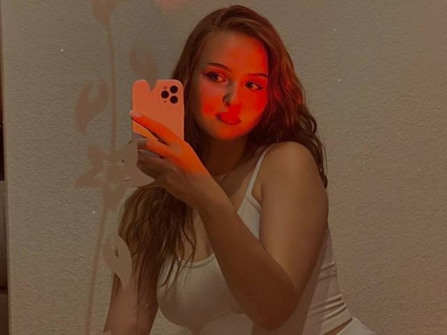 JillianMel, con el rostro iluminado en rojo, sosteniendo un iPhone rosa, vistiendo una camiseta blanca, mostrando sus pechos, y con cabello castaño ondulado en un selfie frente al espejo..