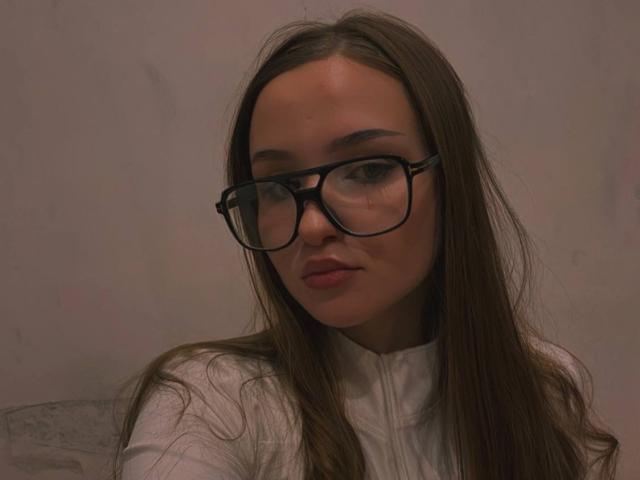 JillianMel with long brown hair, wearing black glasses and a white shirt, has a serious expression and slightly parted lips, looking straight at the camera.
