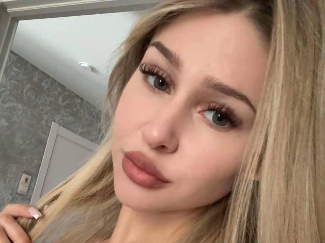 MilagrosEdmisten's face with blonde hair, green eyes, thick eyebrows, long eyelashes, and pink lips. Her hand is near her head. She has a soft, cute look.