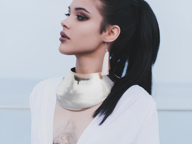MerrillFiler tiene el cabello oscuro y peinado hacia atrás, lleva una blusa blanca, un collar dorado y aretes largos, con un tatuaje debajo del collar, mirando hacia un lado con una expresión seria..