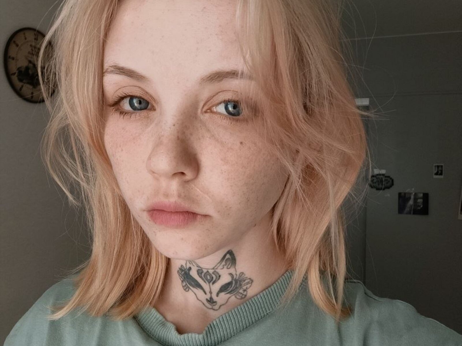 KarissaSuttles has blonde hair, blue eyes, freckles, and a tattoo of a fox on her neck, wearing a green shirt, with a neutral expression.