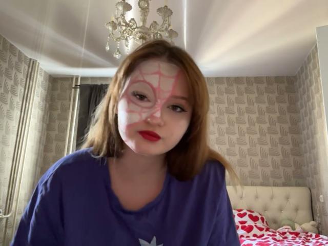 EloisBoeding with painted white face and pink web-like pattern, wearing purple shirt, red lipstick, and brown hair, in bedroom with chandelier and patterned wallpaper.