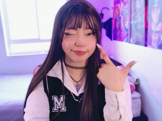 Arieyaara with long brown hair, winking, sticking out tongue, wearing a black vest with an "M", choker, and necklace, pointing up with index finger, in a purple-lit room.