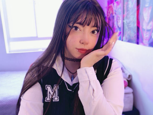 Arieyaara with long brown hair, wearing a black and white school uniform with an "M" on the chest, black choker, and nose ring, touching her cheek with a slight smile.